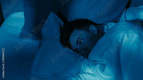 Exhausted man experiencing loneliness insomnia lying awake in bed under dim blue lighting.