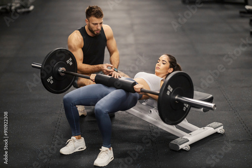 Muscular sportswoman doing hip thrusts on bench with barbell and her coach is motivating her.