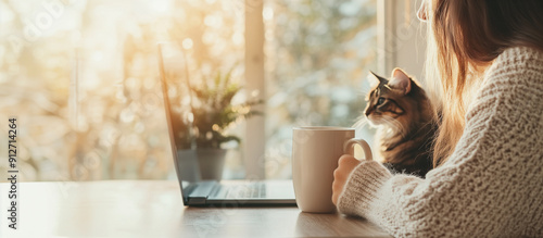 A girl is sitting at a table by the window, holding a mug of tea and looking at a laptop monitor, a cat is sitting next to her and also looking at the monitor. An empty space for the text.