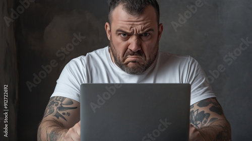 Angry overweight tattooed white man, European looks, sits at a laptop computer. He has a bitter, sulky expression. Landscape format aspect ratio 16:9 with copy space