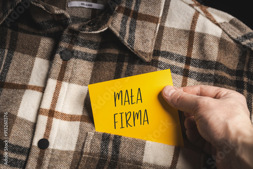 Yellow card with a handwritten inscription "Mała Firma", held in the hand against the background of a brown plaid shirt (selective focus), translation: Small Company