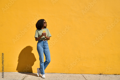 Young happy African gen z girl in sunglasses standing at yellow wall looking at copy space holding smartphone. Smiling Black woman using mobile cell phone advertising ecommerce summer sale. Full body.