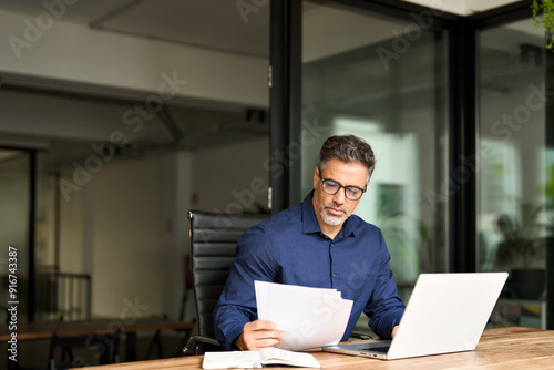 Senior mature male account bank manager executive working on laptop checking paper financial report in office. Busy middle aged professional business man using laptop computer reading document at work