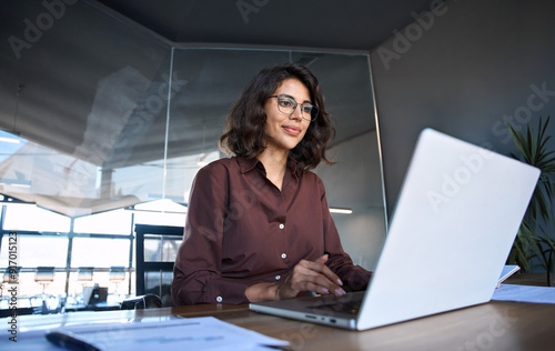 Young professional it specialist latin hispanic business lady working on laptop pc sitting at desk in modern office space. 30s middle eastern indian woman using computer technology app for work online