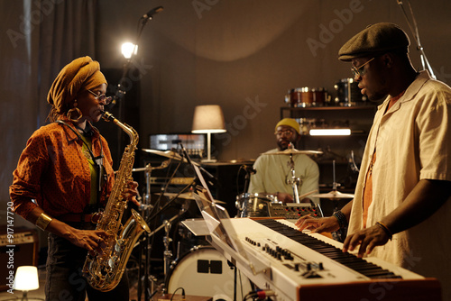 Musicians practicing in intimate studio with saxophone player, keyboardist, and drummer creating lively music. Warm lighting and various instruments adding to the creative ambiance