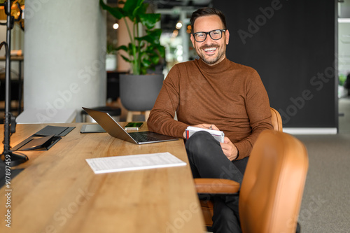 Portrait of inspired male entrepreneur in glasses working over laptop while sitting at desk in office