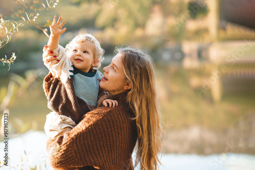 Smiling happy young mother 24-26 year old holding baby girl over lake and nature background. Autumn season. Motherhood.