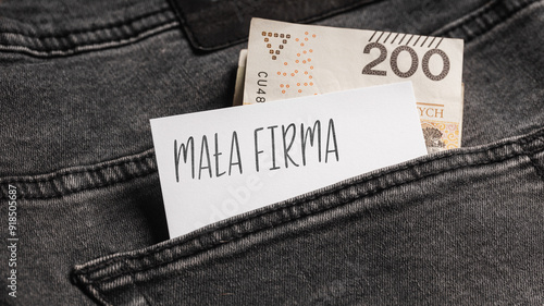 White card with a handwritten inscription "Mała Firma", inserted into the pocket of gray pants jeasnow, next to Polish banknotes PLN (selective focus), translation: Small Company