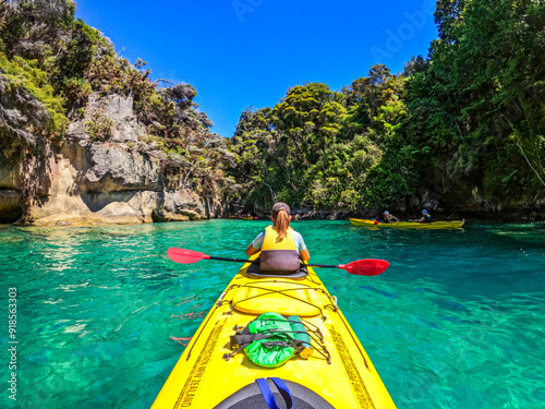 Tourists on a Kayak tour in the Abel Tasman national park. Clear blue turqouise water surrounds the yellow kayak in rocky and bush covered lagoon.