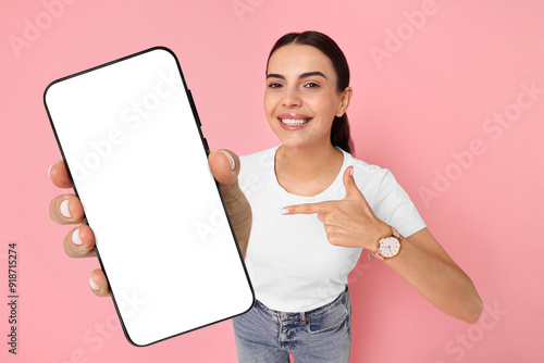 Happy woman pointing at mobile phone with blank screen on pink background. Mockup for design