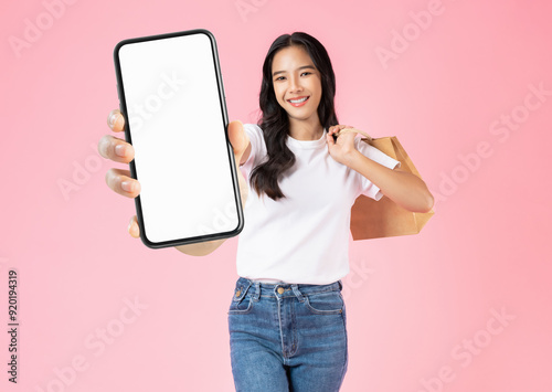 Beautiful Asian woman holding brown blank craft paper shopping bags and showing big smartphone mockup of blank screen on pink background.