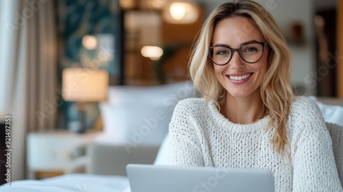 A smiling woman in a white sweater is using her laptop while sitting comfortably in a modern indoor setting, demonstrating a relaxed and productive work atmosphere.