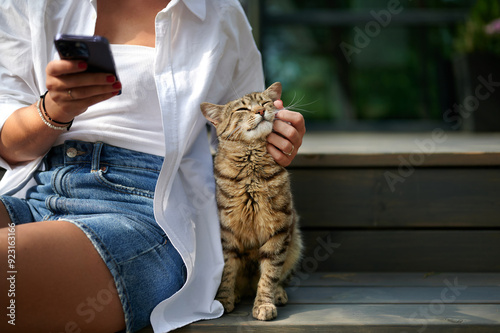 caucasian female sitting on a wooden staircase holding a phone and stroking a beautiful pet cat