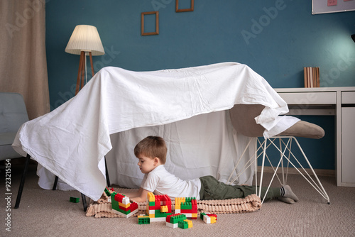 Happy adorable child boy playing in play tent, in handmade toy fort from chairs and blanket