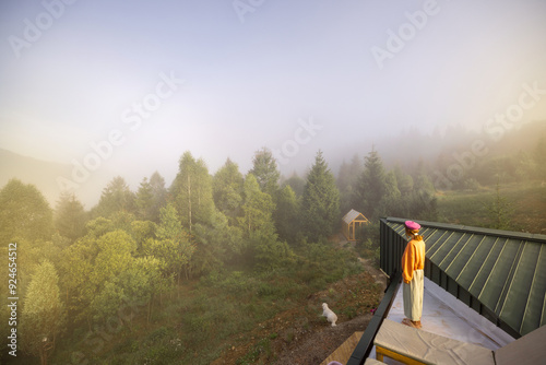 A woman stands on a rooftop, overlooking a misty forest and a small greenhouse, with her dog patiently waiting below, capturing the tranquility of a foggy morning in a secluded natural setting