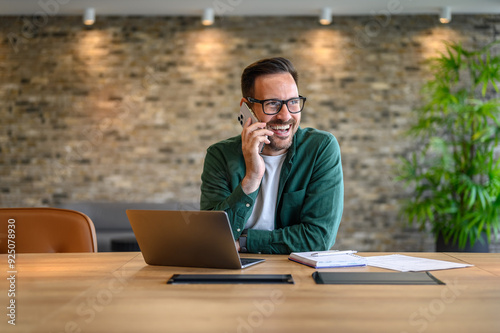 Happy financial advisor in glasses communicating over mobile phone while working over laptop at desk