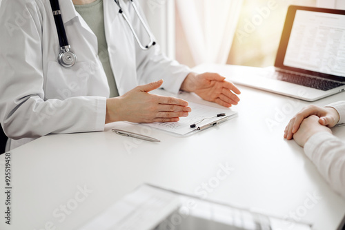 Doctor and patient sitting and discussing something near each other at the white desk in clinic. Female physician is listening filling up a records form. Medicine concept