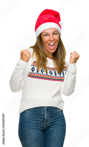 Middle age adult woman wearing winter sweater and chrismat hat over isolated background very happy and excited doing winner gesture with arms raised, smiling and screaming for success