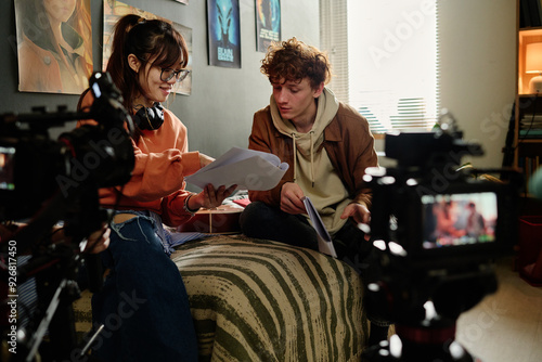 Two young individuals collaborating on a film project in a cozy room filled with cameras and movie posters, one holding script papers while the other listens attentively