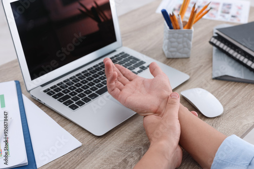 Man suffering from pain in wrist while working on laptop at table indoors, closeup. Carpal tunnel syndrome