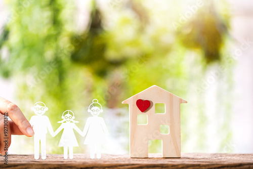 Woman hand holding a family with paper cut to house model and red heart put on the wood on sunlight in the public park, Home sweet home or real estate for love concept.