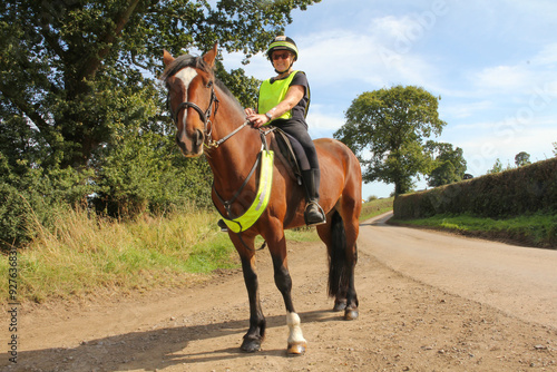 Beautiful bay horse being ridden along country lanes by a woman wearing reflective safety wear, to be seen by other road users and enjoy time together safely in the English countryside.