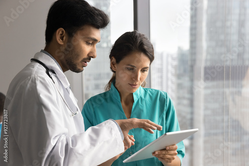 Multiethnic doctors engaged in collaborative medical consultation, discuss patient disease diagnose or treatment plan, reviewing information, focusing on tablet. Teamwork, modern tech in healthcare