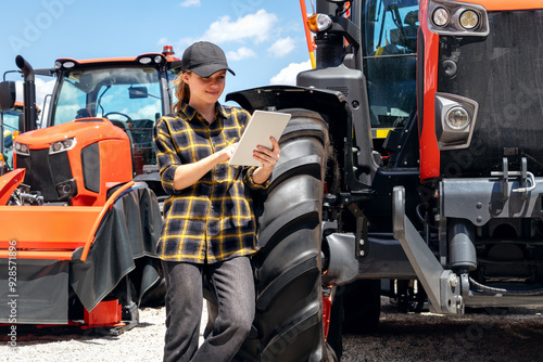 Female sales representative at a dealership for construction and agricultural machinery stands next to a tractor and using digital tablet.