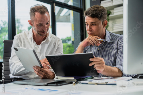 Caucasian middle-aged male businessperson and an Italian accountant are seated at a desk, engaging in a professional meeting, discussing various aspects of technology and innovation in their fields.