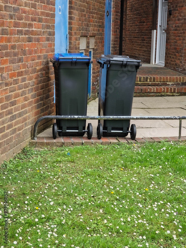 Two black wheelie bins standing on a grassy area next to a brick wall