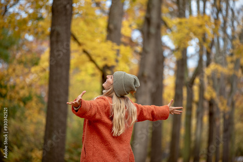 Person in an orange coat and gray beanie stands in a forest with arms outstretched, embracing the autumn season, surrounded by trees with golden leaves, exuding joy and freedom.