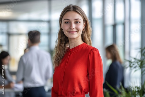 A young woman dressed in a vibrant red blouse smiles confidently in a contemporary office filled with colleagues engaged in work-related activities