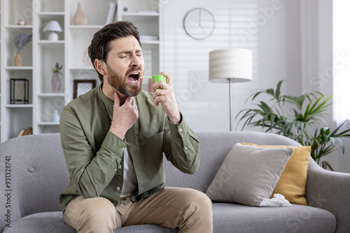 Man sitting on gray couch using throat spray to relieve soreness. Appears to be experiencing discomfort while applying medication. Scene captures moment of self-care in comfortable