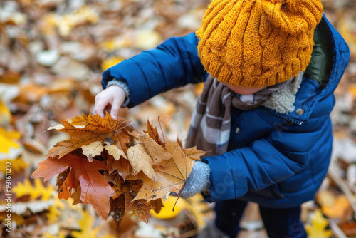 A child in a blue jacket and yellow hat is picking up autumn leaves, a fall season concept.