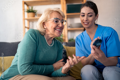 A smiling female nurse in blue scrubs attentively measures the blood glucose level of a happy elderly woman at her cozy apartment, ensuring meticulous in-home patient care.