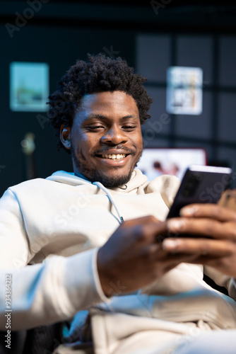 Cheerful man impatiently texting friend, discussing meeting hour. Person sitting on cozy sofa in modern stylish apartment typing on mobile phone, excited to meet up with mate