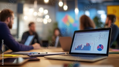 A dynamic office meeting room scene with blurred people engaged in discussion, and a laptop displaying graphs and charts on a wooden table in a modern, creative work space.