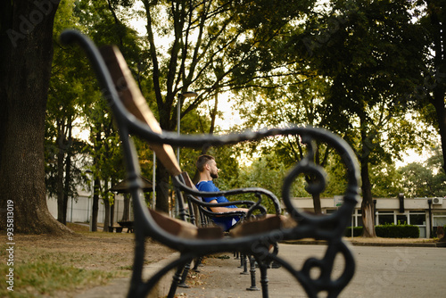 A young bearded male athlete sits and rests after training on a wooden bench in the park. Frame effect, side view. The concept of a sporty and healthy Serbia. Olymp Park in Belgrade.