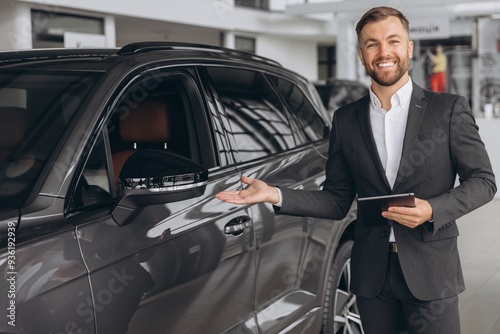 Smiling friendly car seller in suit standing in car salon and holding tablet. It's always pleasure to buy a car on a right place.