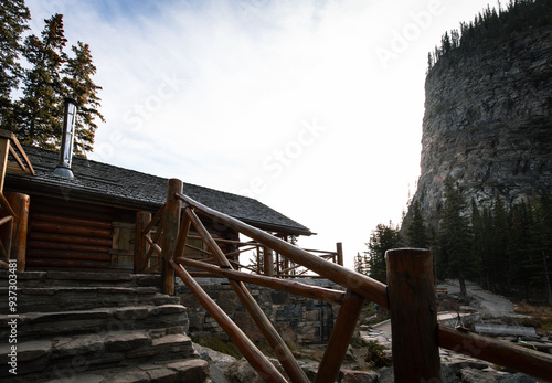 view of a wooden house with stairs in the front with wooden fence in summer with a tall mountain cliff in the background