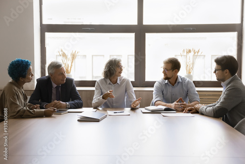 Five young and mature multiethnic staff engaged in briefing at conference table. Shareholders review company financial statement, analyzing profits, losses, revenue, discussing dividend and policies