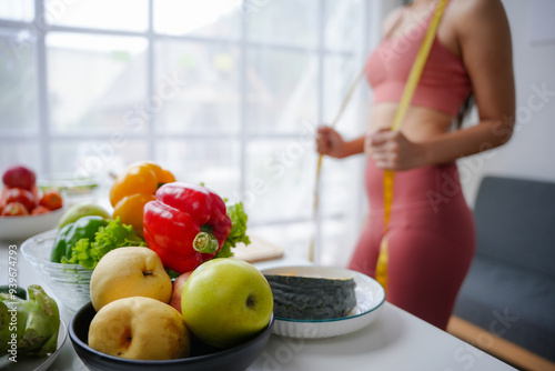 Woman is measuring her waistline in a bright room, with a selection of fresh fruits and vegetables in the foreground