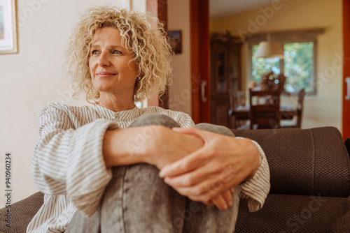 Beautiful smiling relaxed middle aged woman relaxing on sofa at home. Stylish middle aged 50s lady with curly hair, sitting on couch holding a pillow in modern living room.