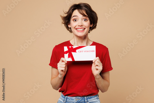 Young shocked surprised happy woman she wear red t-shirt casual clothes hold gift certificate coupon voucher card for store isolated on plain pastel light beige background studio. Lifestyle concept.