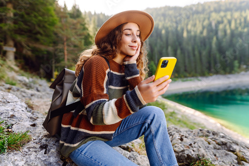 A young woman in a cozy sweater and hat sits on a rock by a beautiful lake, smiling and using her phone. Adventure, nature, blogging.