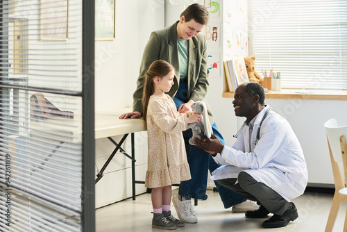 Smiling young girl visiting a pediatric clinic, interacting with a friendly doctor, holding medical equipment while her mother oversees situation