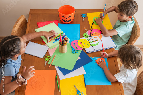 Group Of Children Crafting Halloween Decorations At A Table At Home, Using Colored Paper And Scissors To Create Festive Pumpkin-Themed Art.