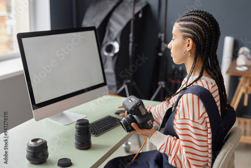 Side view portrait of Black young woman with braided hair as female photographer using computer with blank white screen in photo studio and holding camera