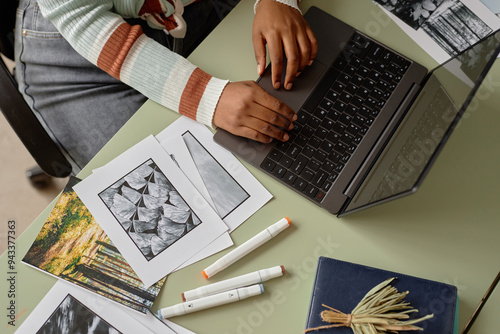 Top view of unrecognizable Black woman using laptop at table with printed photographs and art supplies