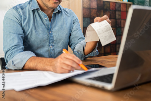 Middle-aged man calculating taxes at a wooden desk with a laptop and holding receipts. Personal finance management and accounting concept during tax season 